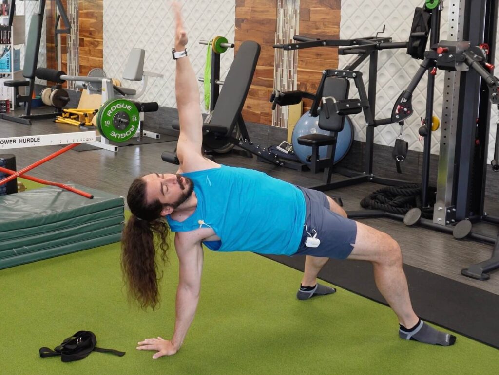 Dr. John extending his hips towards the ceiling supported by both feet and 1 arm with the other arm towards the ceiling for javelin mobility